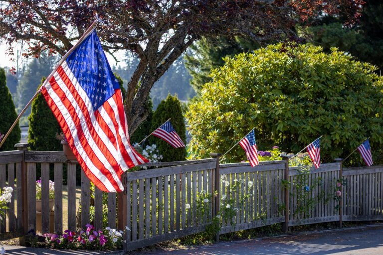 Multiple American flags displayed along a wooden fence with greenery and flowers.