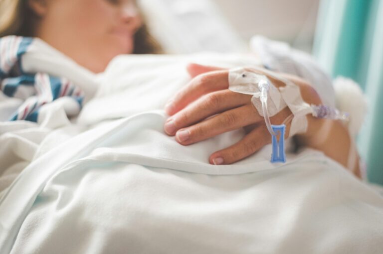 Close-up of a patient’s hand with intravenous catheter, lying in hospital bed.