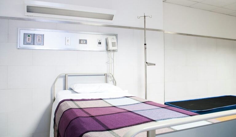 An empty bed in a hospital room with a purple and grey checked blanket.