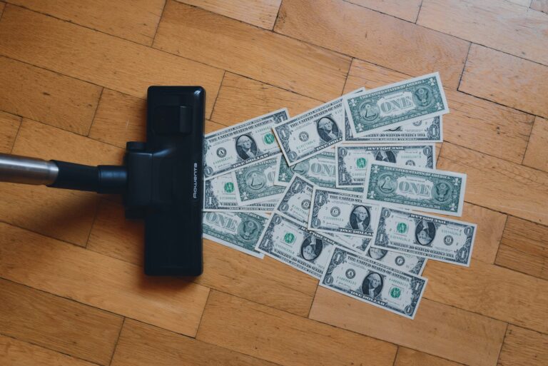 Vacuum cleaner head on wooden floor with scattered US one-dollar bills.