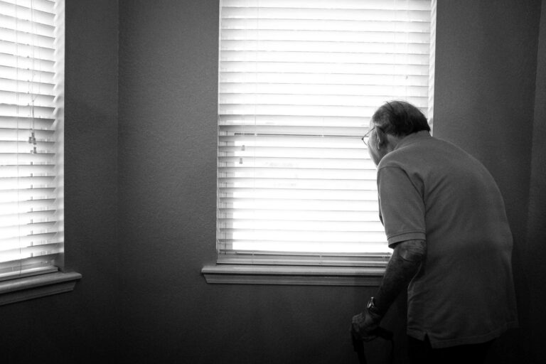 Black and white image of an elderly man with a cane looking out a window with blinds.