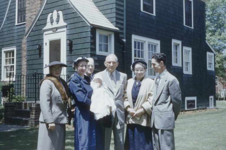 Group of six adults and an infant standing in front of a dark green house with white trim.