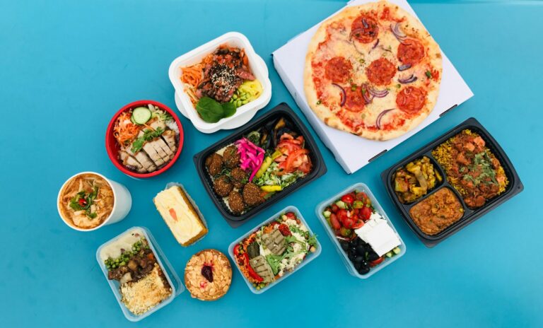 Assorted takeout meals including pizza, salads, falafel, soup, and desserts on a blue surface.