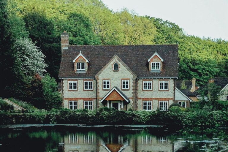 Stone and brick house with white-framed windows, surrounded by greenery, reflected in water.