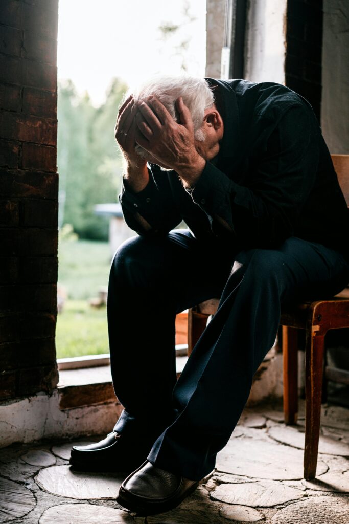 An elderly man sits in a chair with his head in his hands.