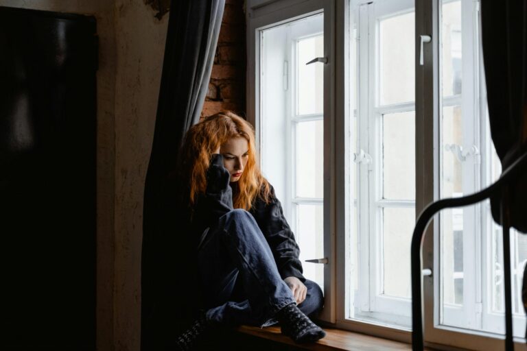 Young woman with red hair sitting pensively on a window sill in dark clothing.