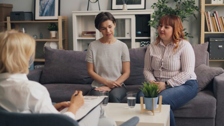 Two women seated on a gray couch in a therapy session with a therapist taking notes.