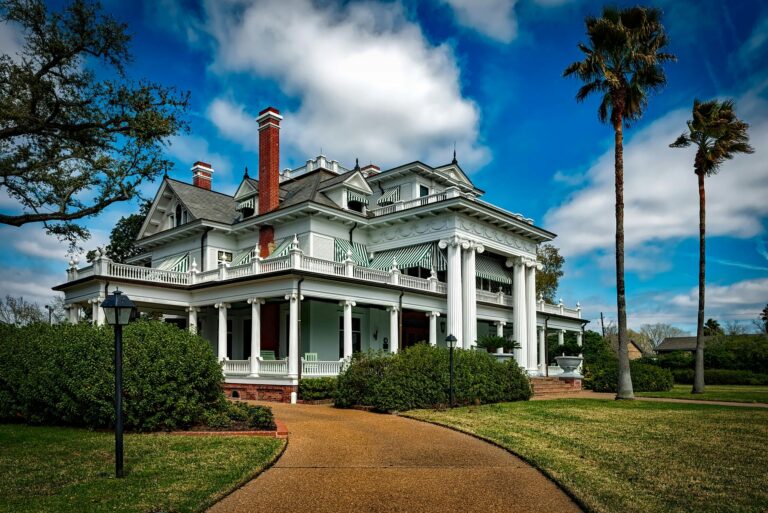 Large white historic mansion with columns, green-striped awnings, and palm trees under a blue sky.