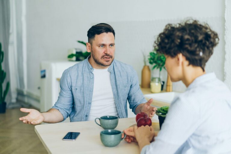 Two men engaged in a serious conversation at a kitchen table with cups and apples.