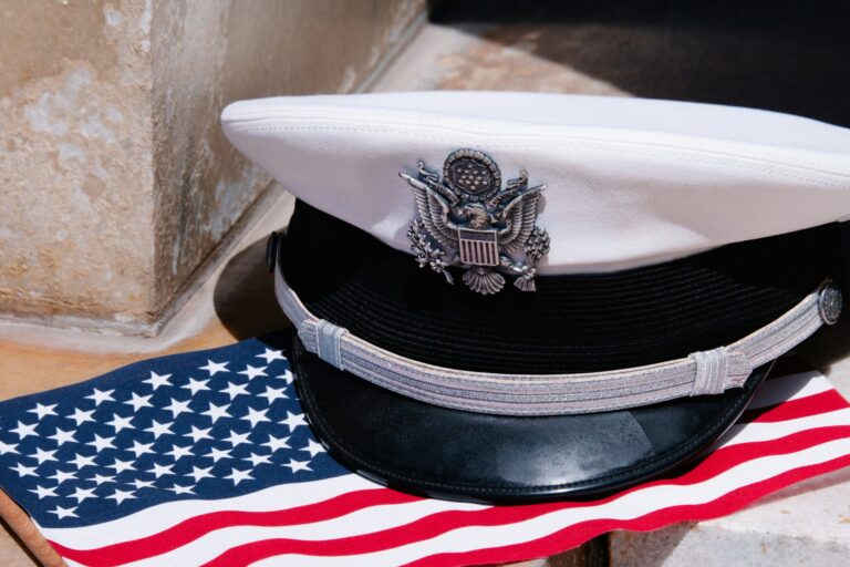 White U.S. Navy officer’s cap resting on an American flag against a stone background.