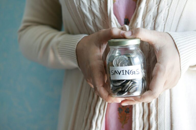 Person wearing a beige sweater holding a glass jar labeled "SAVINGS" filled with coins.