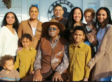 Multigenerational family portrait with nine individuals, including elderly man seated, standing outdoors in front of a house.