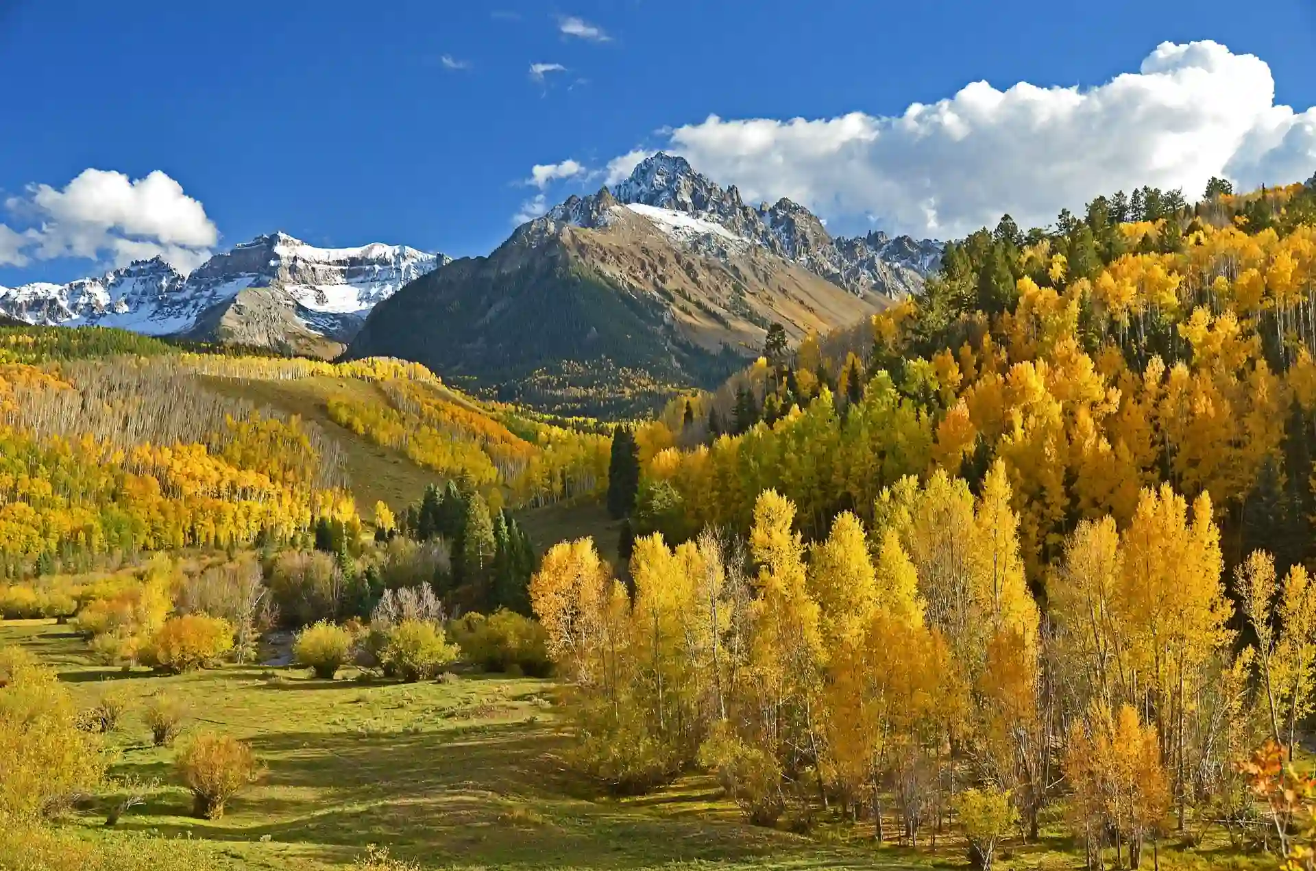 Mountain landscape with autumn-colored trees, green meadow, blue sky, and scattered clouds.