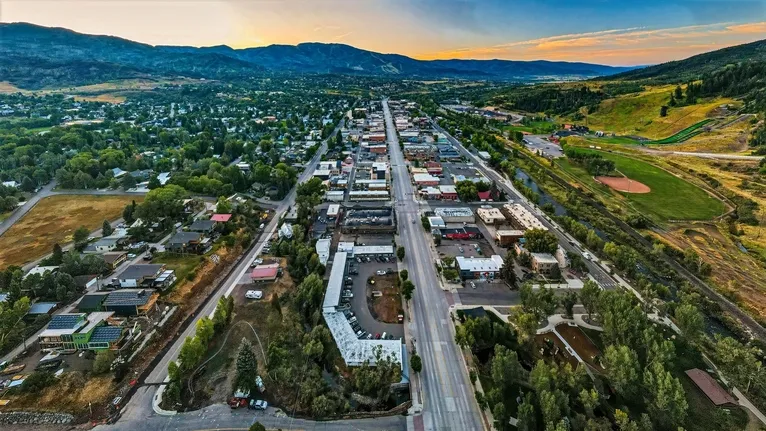 Aerial view of a small town with a central road, surrounding residential areas, greenery, and distant mountains at sunset.