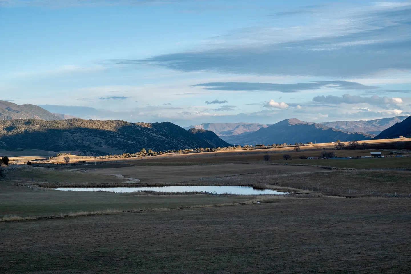 Expansive rural landscape with a small pond, dry grass fields, distant mountains, and a partly cloudy sky.