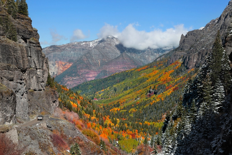 Scenic mountain valley with colorful autumn foliage, rocky cliffs, and a winding road.