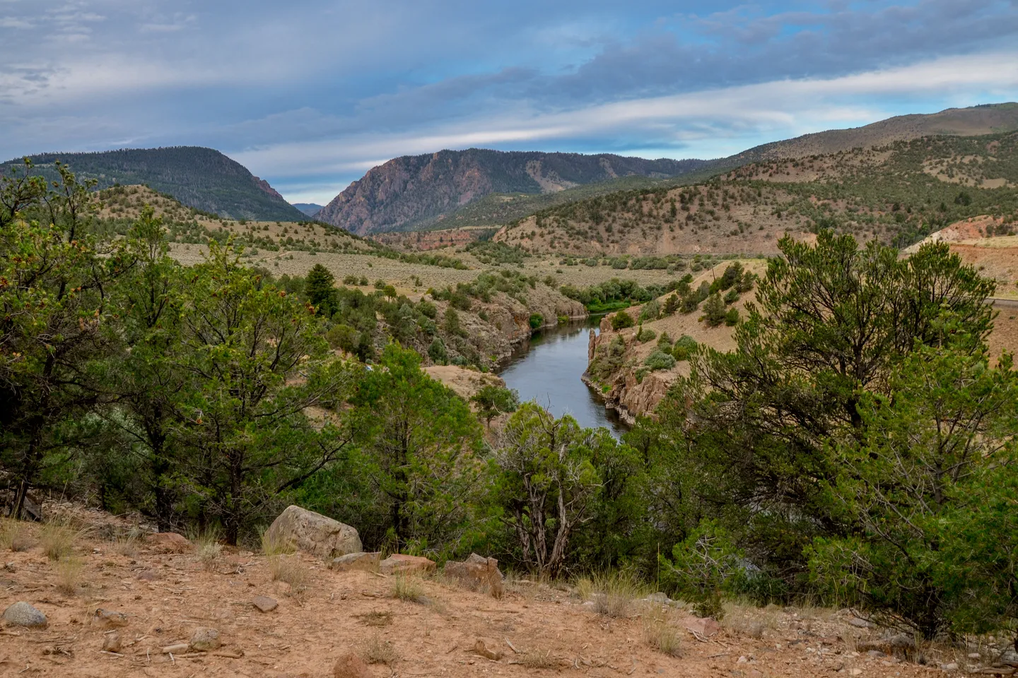 Scenic river winding through arid hills and green pine trees under a cloudy sky.