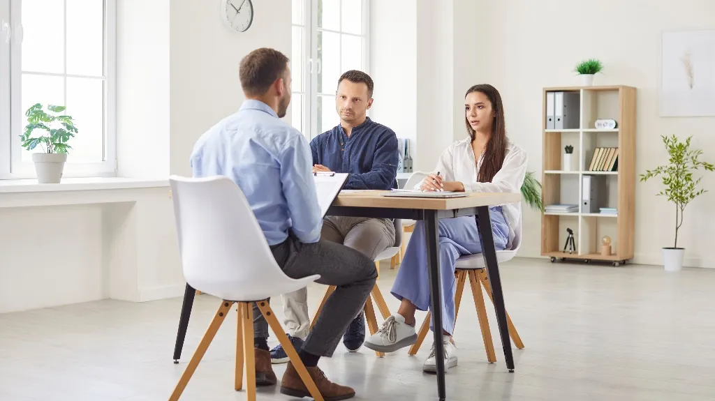 Three professionals engaged in a meeting around a wooden table in a bright, modern office.