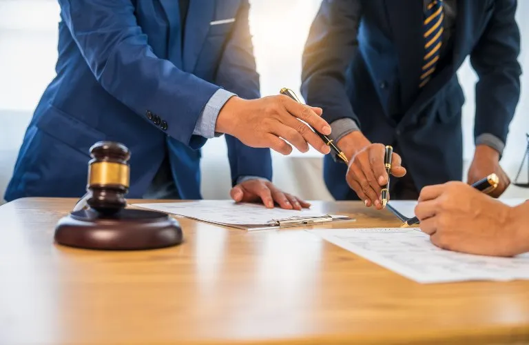 Three individuals in suits signing documents on a wooden table with a judge's gavel nearby.