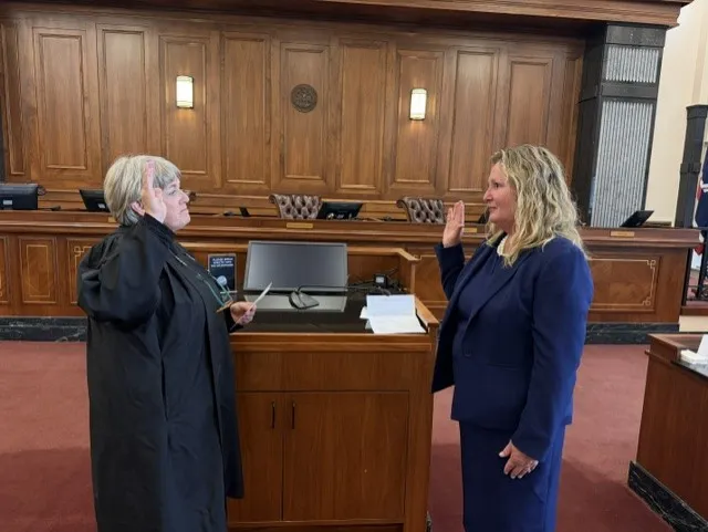 Two women in a courtroom, one in a black robe administering an oath to the other in a blue suit.