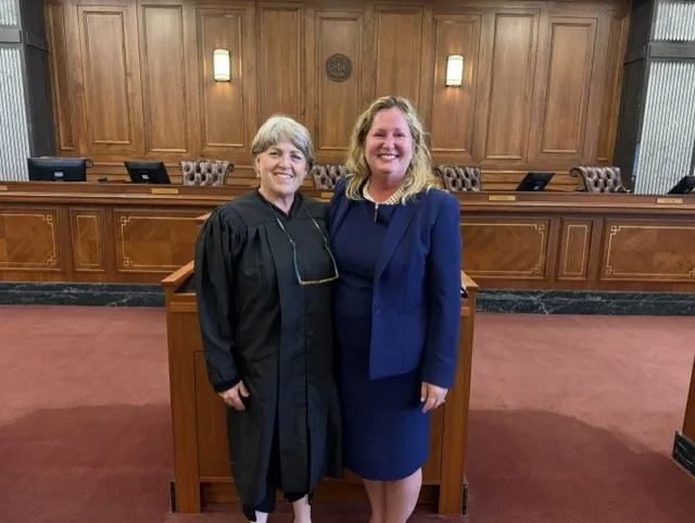Two women standing in a courtroom, one wearing a black judge's robe, the other in a blue suit.