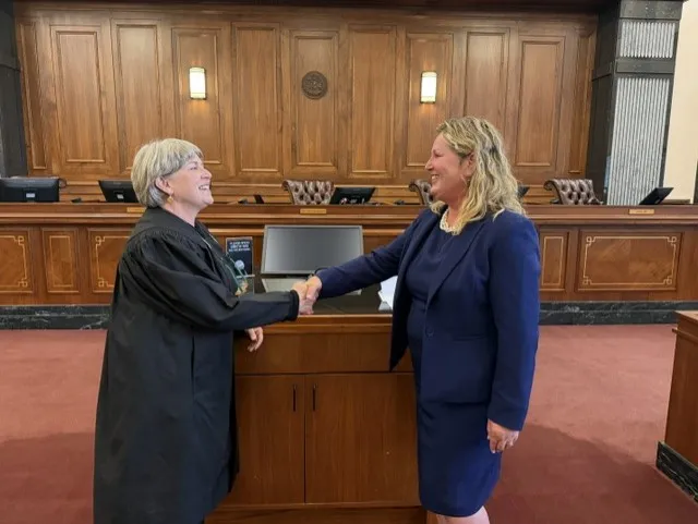Two women shaking hands in a courtroom, one wearing a judge's robe and the other a navy business suit.