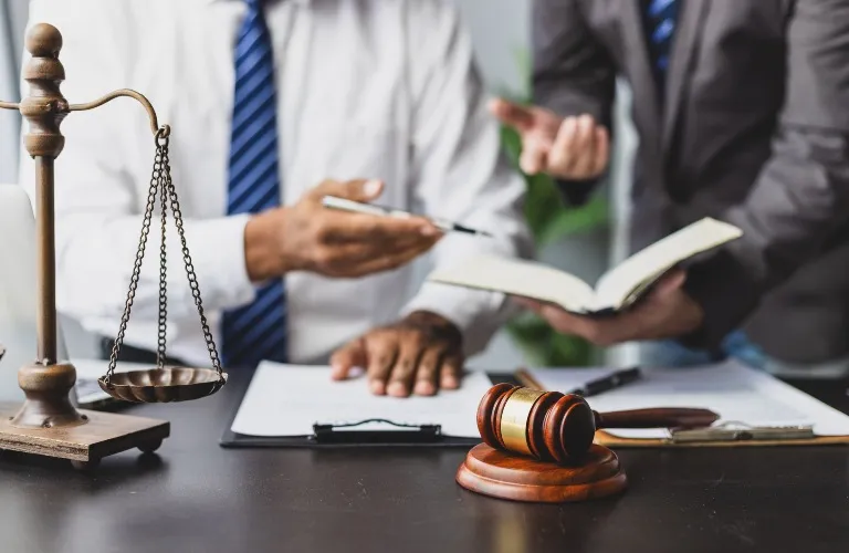 Two professionals in suits discuss a document with legal scales and a wooden gavel on the desk.