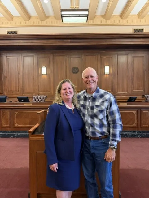 Two individuals, a woman in a navy blue dress and blazer and a man in a blue plaid shirt and jeans, standing and smiling inside a courtroom with wooden paneling.