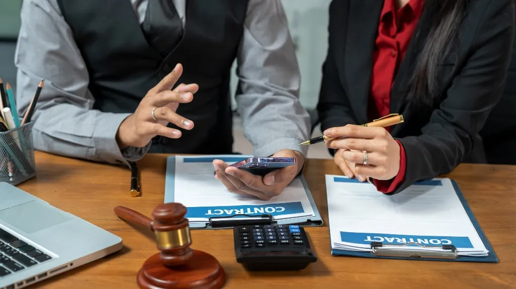 Two professionals reviewing contract documents with a gavel, calculator, and laptop on wooden desk.