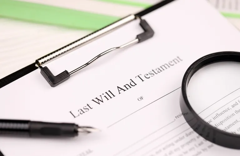 Document titled "Last Will And Testament" on a clipboard with a black pen and magnifying glass.