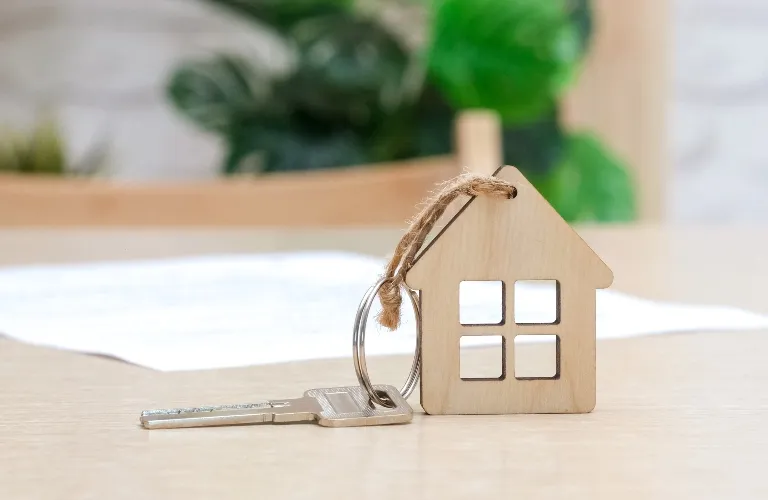 Wooden house-shaped keychain with a metal key on a light wooden surface, blurred background.