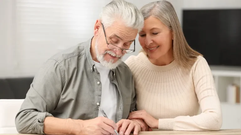Elderly man signing document with elderly woman smiling beside him, indoor setting.