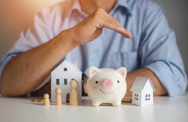 Person in blue shirt gesturing protectively over piggy bank, wooden family figures, and paper houses on table.