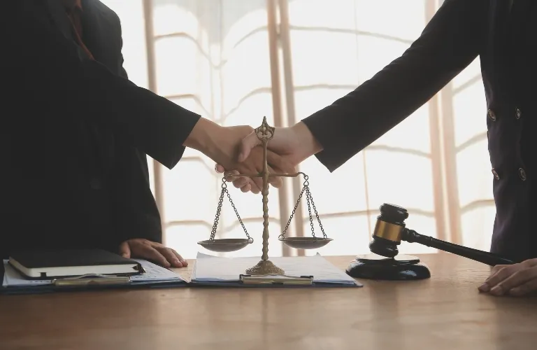 Two individuals in formal attire shaking hands over a desk with scales of justice and a judge's gavel.