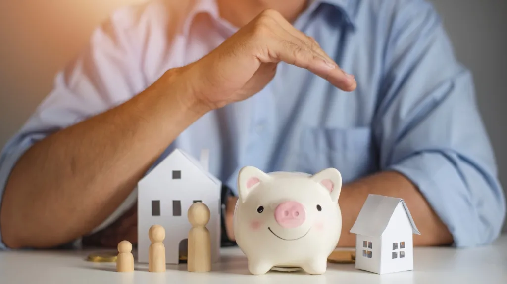 Person in blue shirt protecting white piggy bank with wooden family figures and paper house models.